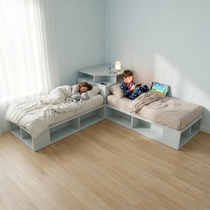 May include: Two white platform beds with built-in storage are arranged in an L-shape. One bed has a person sleeping under a white comforter, and the other has a person using a tablet. A small desk with a lamp and charging station is between the beds.