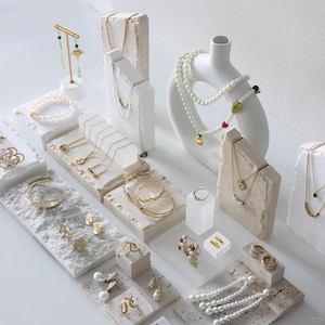 May include: Various gold jewellery items, including necklaces, bracelets, earrings, and rings, presented on textured white plinths. Some pieces incorporate pearls and gemstones. The jewellery is arranged on a white surface.