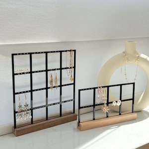 May include: Two black metal jewelry display stands with grid patterns, holding various earrings and necklaces. The stands have wooden bases. A white vase with a necklace is in the background.
