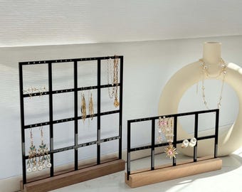 May include: Two black metal jewelry display stands with grid patterns, holding various earrings and necklaces. The stands have wooden bases. A white vase with a necklace is in the background.
