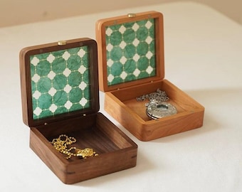 May include: Two wooden jewelry boxes, one open and one closed, sit side-by-side. The boxes have a square shape with a hinged lid. The interior of the lid features a green and white patterned design. One box contains a gold necklace, the other a silver pendant.
