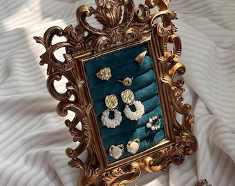 May include: A gold-colored, ornate jewelry display frame with a teal velvet interior. The frame holds various earrings and rings, including pearl and gemstone designs. The frame is set against a white, textured background.
