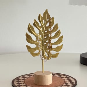 May include: A gold-coloured metal leaf-shaped jewellery holder with a wooden base. Two necklaces, one with pearls and the other with a gold chain, hang from the leaf. The holder sits on a brown and white patterned coaster.