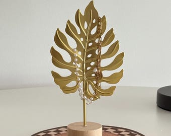 May include: A gold-colored metal leaf-shaped jewelry holder with a wooden base. Two necklaces, one with pearls and the other with a gold chain, hang from the leaf. The holder sits on a brown and white patterned coaster.