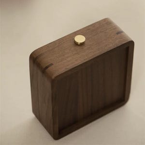 May include: A rectangular wooden box with rounded corners and a brass-colored knob on top. The box is made of dark brown wood, with visible wood grain. The design is minimalist and modern.