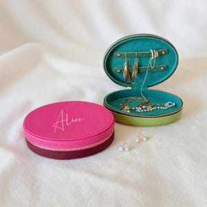 Könnte beinhalten: Zwei ovale Schmuckkästchen, eines geschlossen und eines offen, mit Schmuck. Das geschlossene Etui ist rosa mit dem Namen "Alice" in Schreibschrift. Das offene Etui ist grün mit türkisfarbenem Innenraum und zeigt Halsketten und Ohrringe.