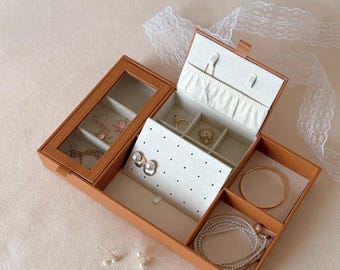 May include: Open, tan leather jewelry box with multiple compartments. The box contains various pieces of jewelry, including earrings, rings, bracelets, and necklaces. The interior is lined with a light-colored fabric and features a clear glass top.