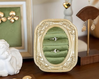 May include: A gold-colored, ornate picture frame with an oval opening, displaying two rings on a green velvet surface. The frame is filled with a green velvet material. Other jewelry items are visible in the background.