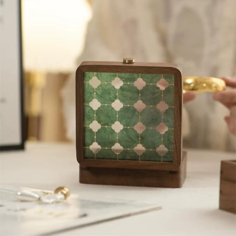 May include: A wooden jewelry box with a green and gold patterned front. The box has a square shape with rounded corners and a small gold knob on top. The box is sitting on a white surface.