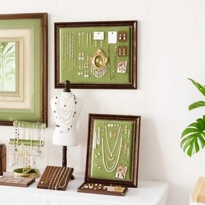 Puede incluir: Una exhibición de joyas, incluyendo collares, pendientes y pulseras, dispuestas sobre una superficie blanca. Las joyas se exhiben en una variedad de soportes y en exhibidores enmarcados con fondos de tela verde. Los marcos tienen bordes de madera marrón oscuro.