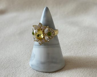 May include: A gold-toned ring with a large yellow oval-shaped center stone, surrounded by smaller white, green, and clear stones. The ring is displayed on a gray and white marble-patterned cone-shaped stand.