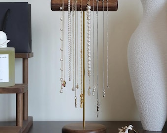 May include: A wooden jewelry display stand with a gold-colored metal pole and base. The stand holds various necklaces with pearls and pendants. The display is on a dark wooden surface, with a white vase and other decorative items in the background.