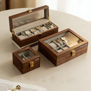 May include: Three wooden watch boxes with glass lids. The largest box holds five watches, the middle box holds four, and the smallest box holds two. The boxes are made of dark wood and have gold-colored clasps. The middle box has "I love you" engraved on it, and the smallest box has "Smith" engraved.