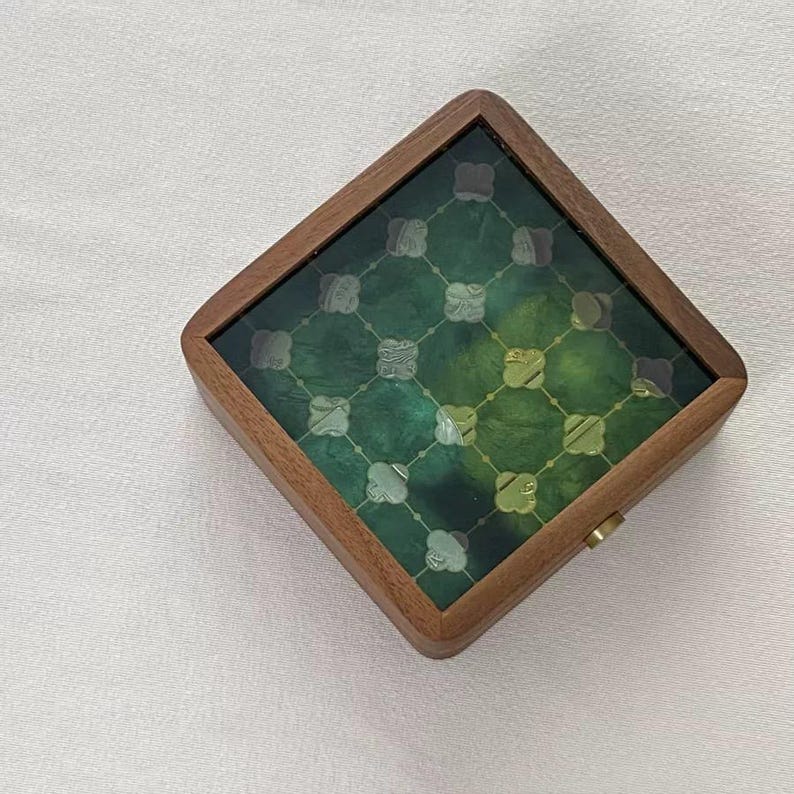 May include: Square wooden box with a green and gold patterned top. The box has a dark wood frame and a decorative design on the glass top. A small gold-colored latch is visible on the side.