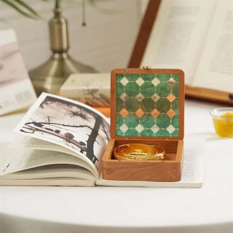 May include: A wooden jewelry box with a green and orange patterned lid, open to reveal two gold bangles. The box sits on a white surface next to an open book with a black and white image.