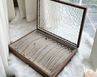 May include: A wooden jewelry box with a clear, textured glass lid. The box is open, revealing several necklaces displayed on a light gray velvet interior. The box is sitting on a white, fluffy surface.