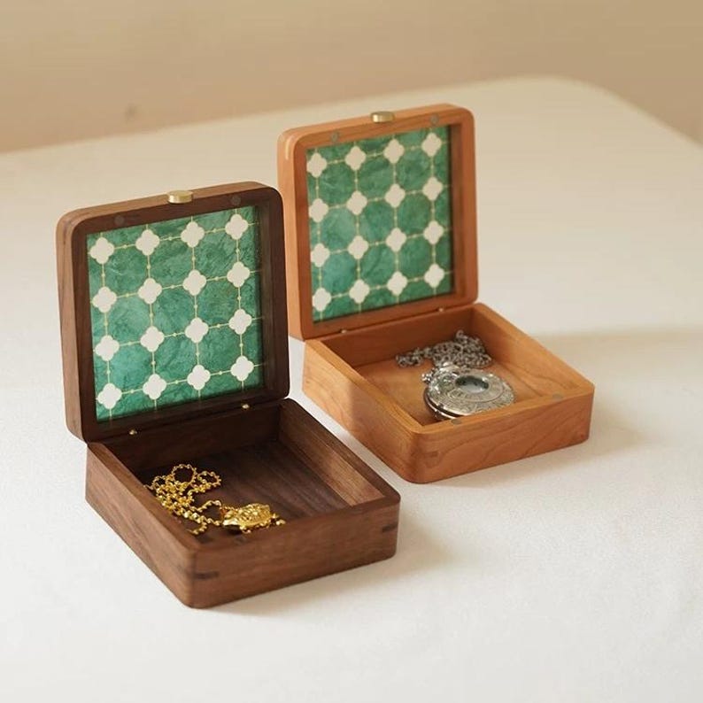 May include: Two wooden jewelry boxes, one open and one closed, sit side-by-side. The boxes have a square shape with a hinged lid. The interior of the lid features a green and white patterned design. One box contains a gold necklace, the other a silver pendant.