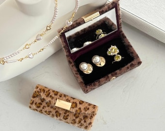 May include: A leopard print jewelry box with a gold clasp, open to reveal a mirror and various rings and earrings. A gold necklace with heart and pearl accents is visible, along with a matching leopard print case.