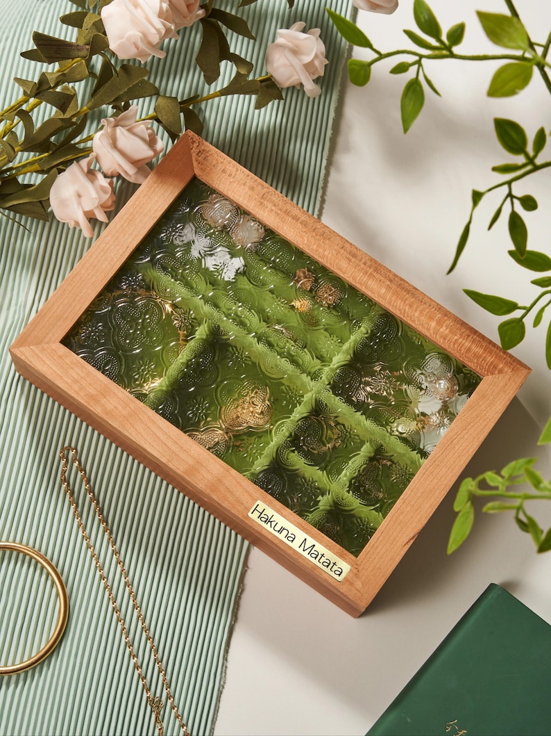May include: A wooden jewelry box with a glass top and green velvet lining. The box has multiple compartments for storing rings, earrings, and other small jewelry items. The box has a gold label that reads "Hakuna Matata".