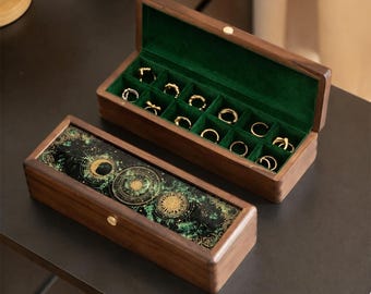 May include: Two rectangular wooden jewelry boxes with dark brown wood grain. One box is open, revealing a green velvet interior with slots for rings. The closed box features a black and green celestial design with gold sun and moon motifs.
