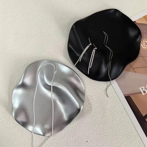 Op de afbeelding: Twee onregelmatig gevormde sieradenbakjes, één zilverkleurig en één zwart, tonen zilveren oorbellen en een ketting. De bakjes liggen op een lichtgekleurd oppervlak en tonen het delicate ontwerp van de sieraden en de contrasterende kleuren.