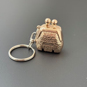 Puede incluir: Llavero de metal dorado en forma de pequeño bolso con superficie texturizada. El llavero tiene un anillo y un cierre. El bolso tiene una pequeña inscripción que dice "CHUANKEFANG".