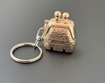May include: Gold-toned metal keychain shaped like a small purse with a textured surface. The keychain has a ring and a clasp. The purse has a small inscription that reads "CHUANKEFANG".