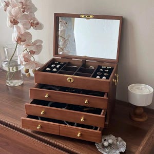 May include: A wooden jewellery box with a mirrored lid and multiple drawers. The box is open, revealing compartments for rings and other jewellery. The drawers are lined with black velvet and have gold-coloured knobs. The box is on a wooden surface.