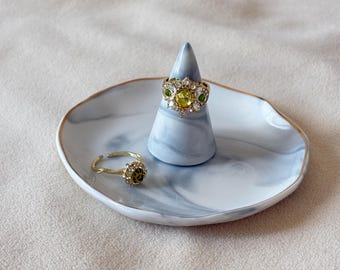 May include: A marble-patterned ring dish with a gold rim holds two rings. One ring is on a blue and white cone-shaped ring holder. The rings are gold-toned with green and clear gemstones. The dish is white with blue marbling.