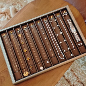 May include: A jewelry display tray filled with various gold necklaces, earrings, and a wristwatch. The necklaces feature different designs, including chains, clover motifs, and pearl accents. The watch has a silver band and a rectangular face. The tray is brown with a silver frame.