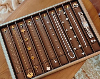 May include: A jewelry display tray filled with various gold necklaces, earrings, and a wristwatch. The necklaces feature different designs, including chains, clover motifs, and pearl accents. The watch has a silver band and a rectangular face. The tray is brown with a silver frame.