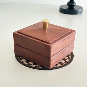 Könnte beinhalten: Kleine, quadratische Holzbox mit rotbraunem Finish und einem goldfarbenen Knopf auf dem Deckel. Die Box steht auf einem runden Untersetzer mit braunem und weißem Schachbrettmuster. Aus Holz gefertigt.