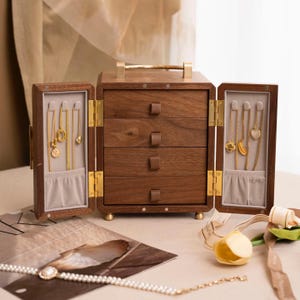May include: A wooden jewelry box with four drawers and two side compartments. The box is made of dark wood with brass hardware and a gold handle. The side compartments display gold necklaces. A pearl necklace and other jewelry are on the table.