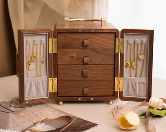 May include: A wooden jewelry box with four drawers and two side compartments. The box is made of dark wood with brass hardware and a gold handle. The side compartments display gold necklaces. A pearl necklace and other jewelry are on the table.