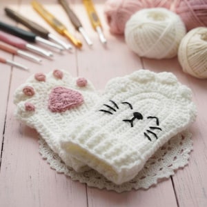 Könnte beinhalten: Weiße gehäkelte Katzenpfoten-Fäustlinge mit rosa Pfotenpolstern und einem herzförmigen rosa Pfotenabdruck. Die Fäustlinge haben ein Katzengesicht-Design mit schwarzen Stickereidetails. Häkelnadeln und Wollknäuel im Hintergrund.
