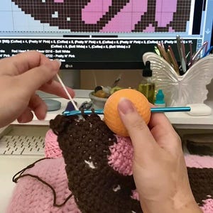 May include: A blue crochet hook with a round orange ergonomic handle is being used to work on a dark brown and light pink crocheted fabric. The project is guided by a digital pattern displayed on a computer monitor in the background.