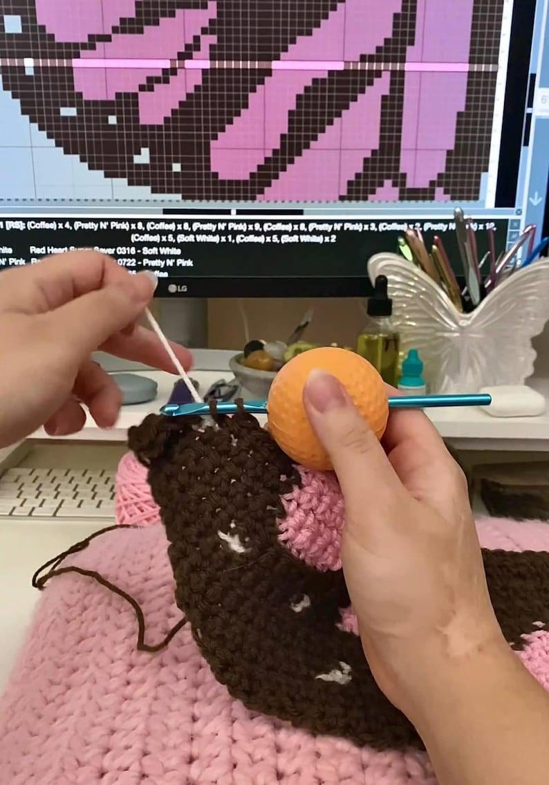 May include: A blue crochet hook with a round orange ergonomic handle is being used to work on a dark brown and light pink crocheted fabric. The project is guided by a digital pattern displayed on a computer monitor in the background.