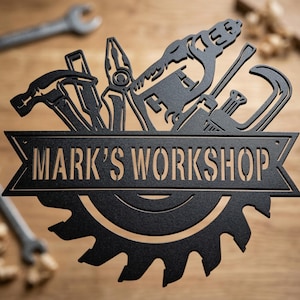 May include: Black metal sign with the text "MARK'S WORKSHOP" above a saw blade design. Silhouettes of tools, including a hammer, pliers, and wrench, are arranged above the text. The sign is set against a wooden background.