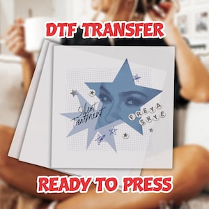 May include: A stack of white DTF transfer sheets. The top sheet features a blue starburst design with the text "Silent Treatment" and "Freya Skye". The design includes a close-up of eyes and small star illustrations. The words "DTF TRANSFER" and "READY TO PRESS" are in red.