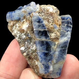 May include: A raw mineral specimen featuring deep blue, translucent crystalline structures embedded within a textured, brownish-tan matrix. The stone displays distinct vertical striations and a jagged, uneven surface with a mix of opaque and semi-transparent sections.