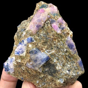 May include: A raw mineral specimen featuring embedded crystalline structures in shades of deep blue, violet, and soft pink. The host rock is a textured, brownish-grey matrix with a rough, granular surface that contrasts with the smooth, translucent facets.