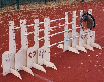 May include: Wooden horse jump set with six jumps. Each jump has a unique design, including horse silhouettes and heart cutouts. The crossbars are painted in various colors. A stuffed horse head is attached to the last jump.