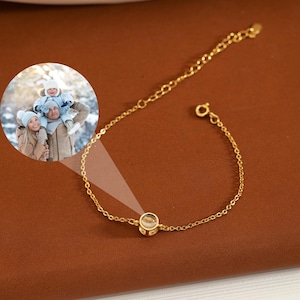 Puede incluir: Una delicada pulsera dorada con una pequeña gema redonda y transparente en el centro. La pulsera está sobre una superficie marrón. Una imagen circular de una familia está en la esquina superior izquierda.
