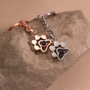 Può includere: Due portachiavi a forma di fiore, uno oro rosa e uno argento, ciascuno con un piccolo centro nero. I portachiavi sono su una superficie di pietra. Il portachiavi oro rosa è a sinistra e quello argento a destra.