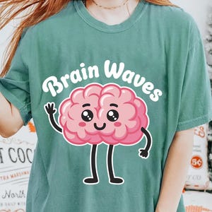 May include: Teal-colored t-shirt featuring a cartoon brain with a smiling face, arms, and legs. The text "Brain Waves" is above the brain graphic in a playful font. The shirt is a comfortable, casual style.