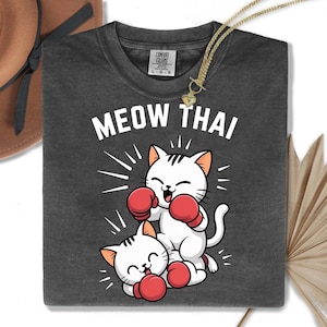 Puede incluir: Camiseta gris oscuro con el texto "MEOW THAI" y un gráfico de dos gatos blancos con guantes de boxeo rojos. Los gatos están en posición de combate. Un collar dorado está sobre la camiseta. Marca Comfort Colors.