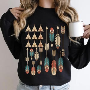 May include: Black sweatshirt featuring a Native American-inspired design. The design includes teepee shapes, arrows, and feathers in shades of teal, orange, and beige. The sweatshirt is a crew neck style.