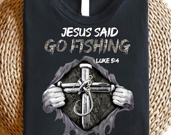 Peut inclure: T-shirt noir avec l'inscription "JESUS SAID GO FISHING" et "LUKE 5:4". Le motif représente une croix faite de clous avec un hameçon, donnant l'impression de déchirer le t-shirt.