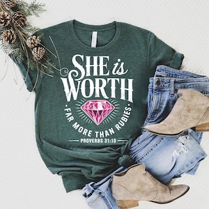 May include: Dark green t-shirt with the text "SHE IS WORTH FAR MORE THAN RUBIES" and a pink diamond graphic. The shirt is paired with blue jeans and tan ankle boots. A sprig of pine and pinecones is in the upper left corner.