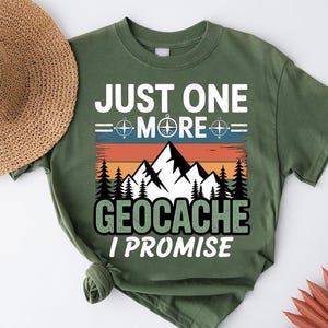 May include: Olive green t-shirt with the text "JUST ONE MORE GEOCACHE I PROMISE" in white and black. The design includes a compass, mountains, and trees, with a sunset-colored background. A straw hat is visible in the background.